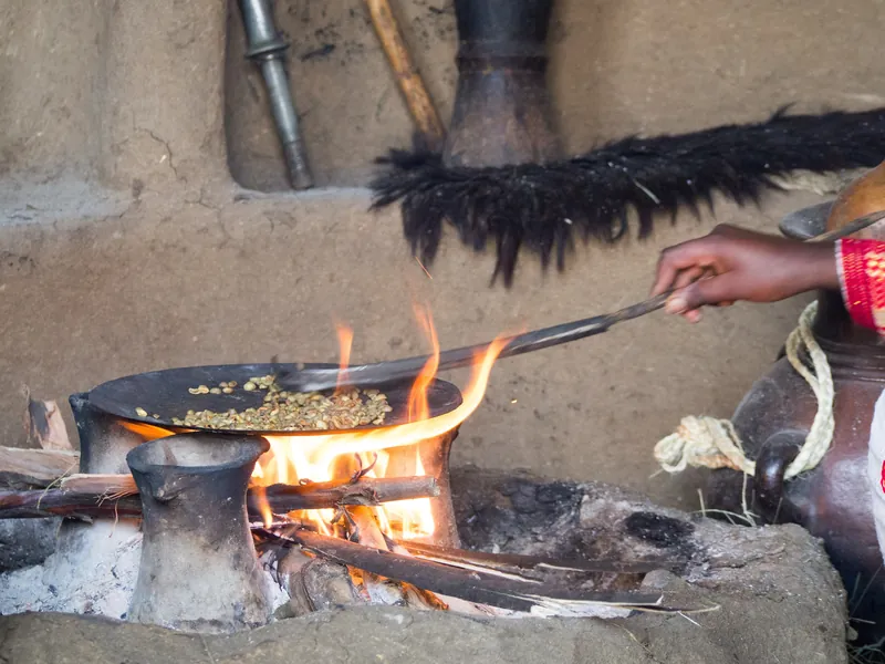 Hands roasting coffee beans over open flames next to a clay jebena